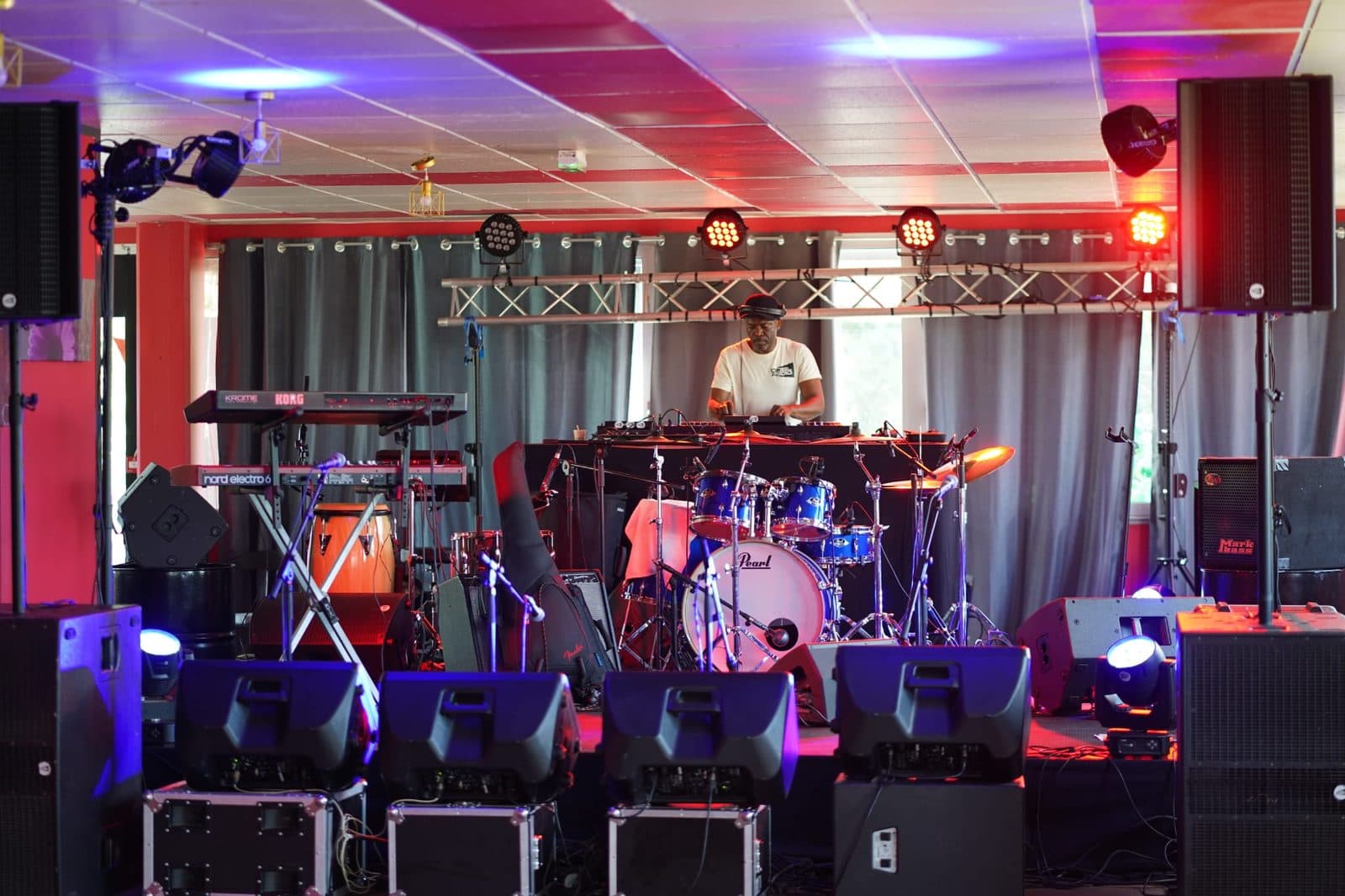 Plateau de concert équipé d'une batterie, de claviers, de retours de scène et d'un pont lumière allumé en rouge et bleu.